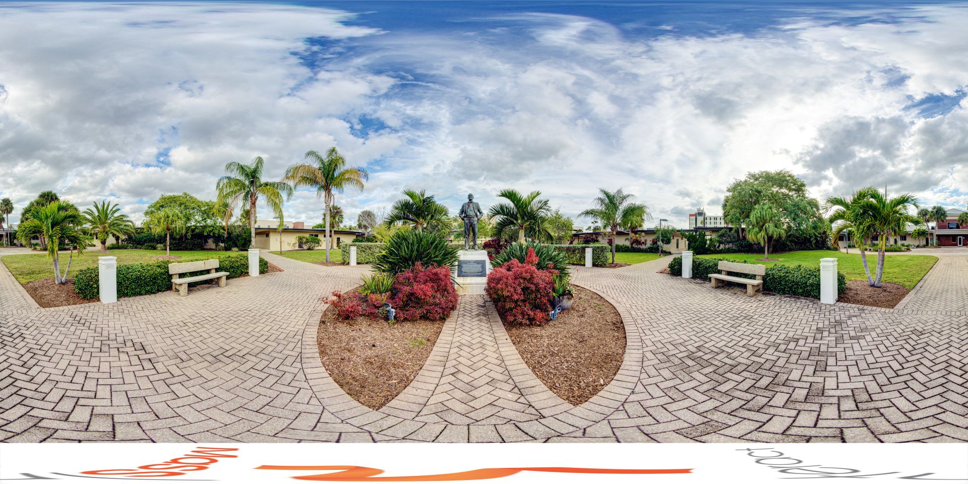 Panoramic view of a lush academic quad featuring a statue of Jerome P. Keuper, palm trees, benches, and brick pathways under a cloudy sky